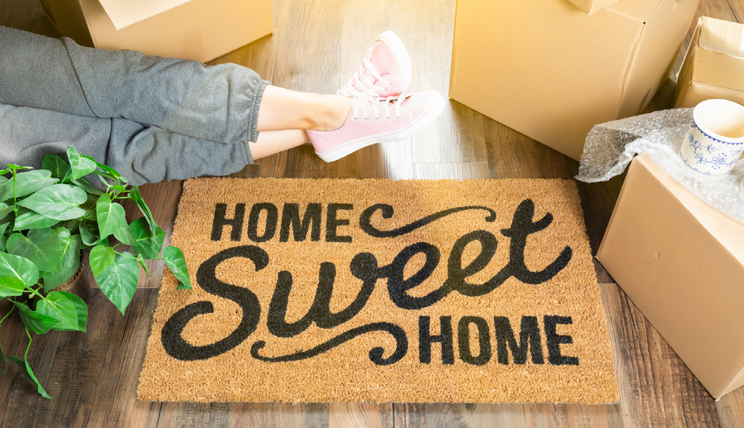 woman wearing sweats relxaxing near home sweet home welcome mat, moving boxes and plant